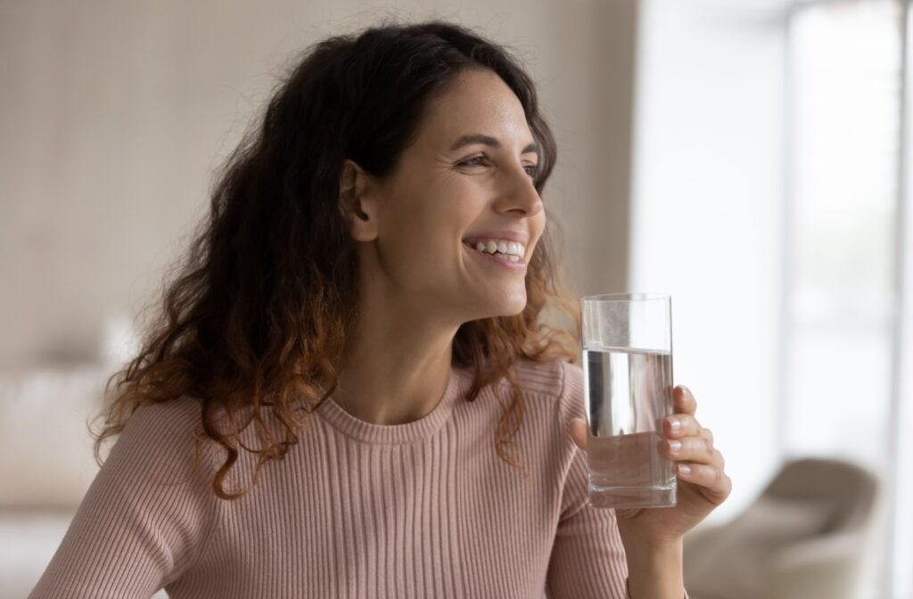 Woman in a pink ribbed shirt drinking fluoridated tap water at home smiling.